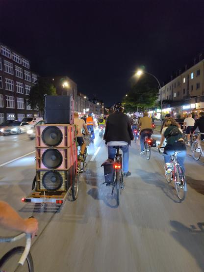 Ein Fahrrad mit Lastenanhänger, der mit einem nach hinten gerichteten Turm aus Lautsprechern bestückt ist. Daneben mehrere Radfahrende, die zusammen an der Critical Mass in Hamburg teilnehmen.