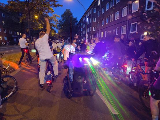 Ein Fahrrad mit Lastenanhänger, der mit nach hinten gerichteten Lautsprechern und Lasershow und Nebelmaschine bestückt ist. Daneben mehrere Radfahrende, die zusammen an der Critical Mass in Hamburg teilnehmen.