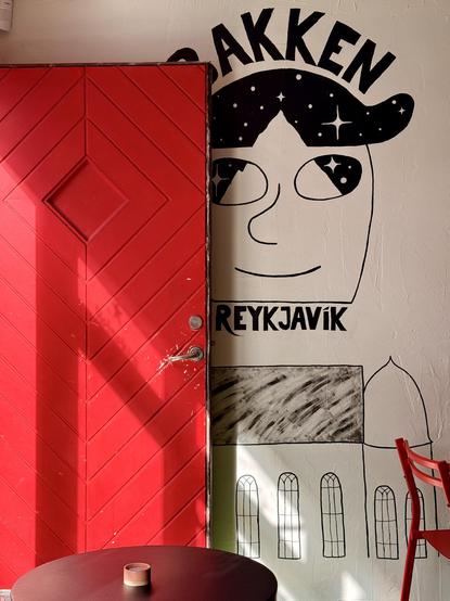 A red door lets streaming sun come into the bar. There’s a mural on the wall behind the door and it has the name of bar on it (Bakken)
