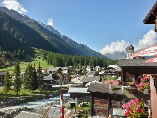 Links grüne Wiesen und Wald am Berghang, mittig und rechts das Dorf Blatten, im Borgenlicht, in der Ferne der Kirchturm und die Berge des Tal-Endes
