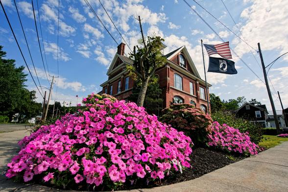 A huge clump of petunias mostly fills the lower left 1/4 of the frame,  a similar clump can be seen further away and behind both of those a flagpole with an American and POW/MIA flag and a small historic brick building with a square footprint seen corner-on.  Behind it all a blue sky with high-altitude clouds with power lines that don't quite meet at a 90 degree angle because I used a wide angle lens and I was looking up from the ground.