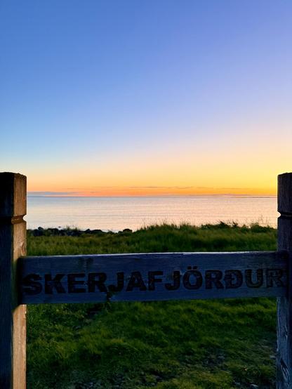 A park sign saying “Skerjafjörður” is in the foreground with lush green grass and a vivid sunset in the background. 