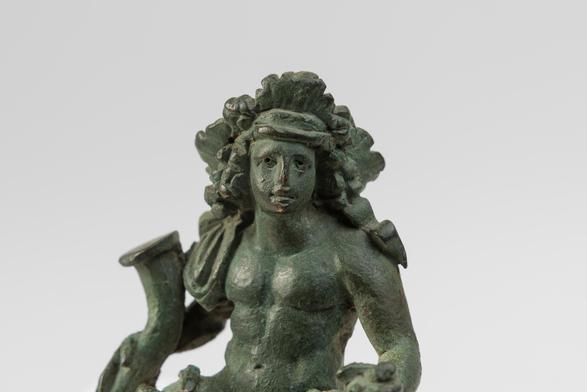 This bronze sculpture is an exquisitely detailed representation of the god Dionysos, whose cult was one of the most popular in ancient times. Here, the god is nude, with a mantle that gently lies on his right leg. He is holding a bunch of grapes with his left hand, and a rhyton (drinking horn) with the right. The god is youthful and joyfully gazes forwards, with a crown of ivy leaves set upon his long flowing hair.

These features clearly resemble those of the life-size Roman marble sculpture o…