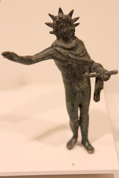 Bronze figurine of the Roman sun god Sol. He's standing, his right arm raised, a riding crop in his left. A seven-spiked sun ray crown adorns his head. A himation is draped around his shoulders and left arm.