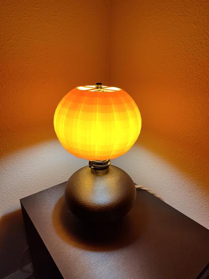 Lamp on table, unlit: A handmade lamp with a pumpkin-shaped orange shade resting on a shimmering black metallic base, sitting on a wooden table.