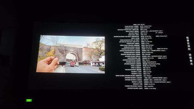 영화 <난징사진관> 엔딩크레딧이 극장 은막에 상영되고 있다. 오른쪽에는 출연진 이름들이 있고, 왼쪽에는 남경대학살 당시 사진을 사진 속 배경의 장소에서 들고 서 있는 화면이 보인다.