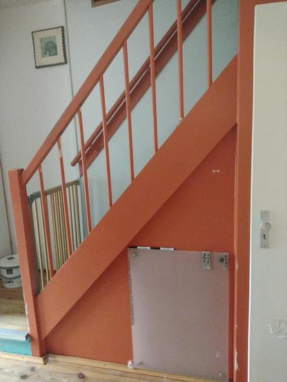Foto van een hal met trap. De trap, leuning en houtwerk eronder is oranje, deur en muren zijn verschillende tinten wit.