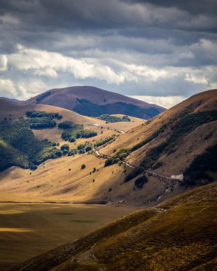 Rolling hills and mountains south of Castelluccio di Norcia, Italy, with a winding road cutting through grassy and forested slopes under a partly cloudy sky.