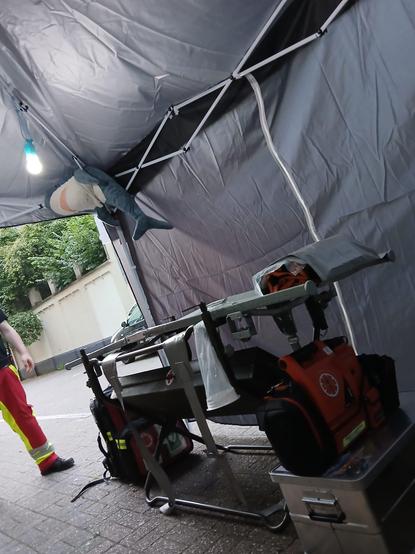 Auf dem Bild sieht man nochmal das Tragelagerungsgestell (dadrauf kann man eine Trage legen sodass man Rückenschonend auf Höhe arbeiten kann), unter dem Kopfteil der Trage steht eine Kiste, in dieser haben wir alles was wir so brauchen für das Zelt, Laken, lampen, Handschuhe etc. dadrauf steht unser 08/16 EKG Defibrillator. auf der Beinseite der Trage steht unser Notfallrucksack mit Defibrillator und Sauerstoff für unseren Fuß Trupp für unterwegs. an der decke des Pavillions Hängt Haidi und guckt von oben auf die Trage und eine Lampe.