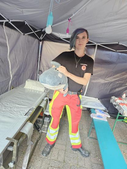 Eine unserer Sanitäterinnen steht in der Erstehilfe Station mit einem Ikea Hai (unser Maskottchen Haidi) die Sanitätsstation ist in einem Pavillion dort steht eine Trage auf der Linken Seite mit Tragenlagerungsgestell, hinten an der wand ein Feltbett und Rechts eine Bierbank und Tisch mit Medizinischem Material drauf 