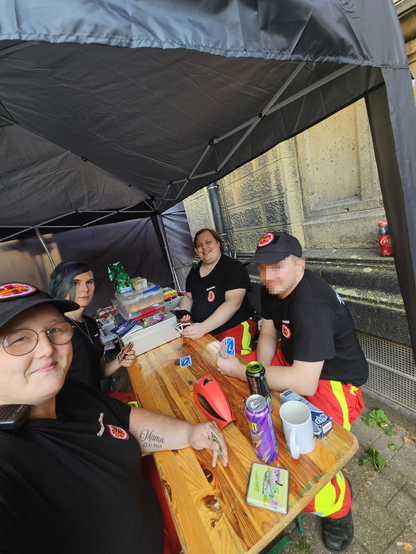 4 Demosanitäter*innen sitzen zusammen auf der Bierbank und dem Tisch und spielen zusammen Uno, alle sind gut gelaunt und haben spaß.