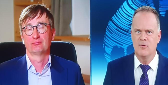 Das Bild zeigt zwei Männer im Heute Journal. Der Mann auf der linken Seite heißt Prof Neitzel und trägt einen dunkelblauen Anzug mit einem hellblauen Hemd und hat kurzes, braunes Haar. Er sitzt in einem Raum mit einem hellen Hintergrund und einem Teil eines Regals im Hintergrund. Der Mann auf der rechten Seite trägt einen dunkelblauen Anzug mit einem weißen Hemd und einem dunkelblauen Krawatten mit Punkten. Er steht vor einem blauen Hintergrund mit einem abstrakten, kreisförmigen Muster, das an…