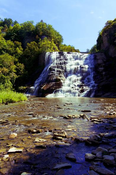 Roughly the bottom half is a shallow but wide creek with a few rocks visible above the water and others visible below the water,  a little bit of a vegetated bank comes in at the middle left.  The main event is a tall waterfall of brown rock with a large number of cascades on it;  there are a lot of trees to the left,  with a fall kind of green that was real,  not just color graded,  and an almost perfectly clear blue sky above it.