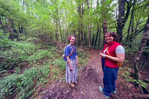 Two woman look small relativet to a big forest,  one near the center has her hair in a red kerchief and wears a blue blouse and long blue and white pleated skirt,  another halfway to the left has ringlets and is stretched out horizontally by the ewide angle lens and has a red vest and jeans.  They are standing on a dirt trail and there is a patch of small vegetation to the left but the to the right and the rear tall trees.