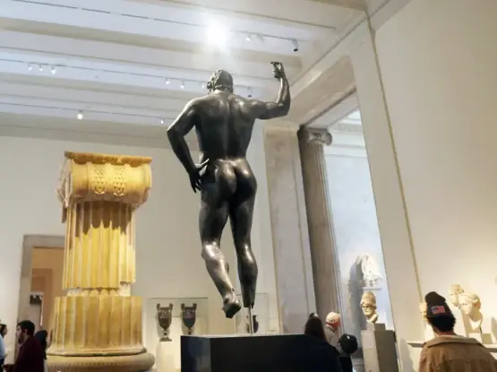 Ancient Greek bronze sculpture of a male figure, seen from behind. He holds one arm up as if he was holding a staff or spear that is now missing, his other hand resting on his hips. His glutes are exquisite.