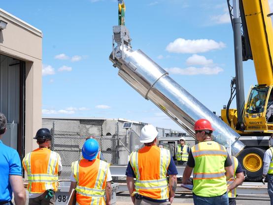 Four hard hat workers wearing high-viz clothes look on as a crane lifts a stainless steel cylinder,  now at a 90 degree angle to transfer into a building which can be barely seen on the left;  behind it a blue sky with a few cumulus clouds.