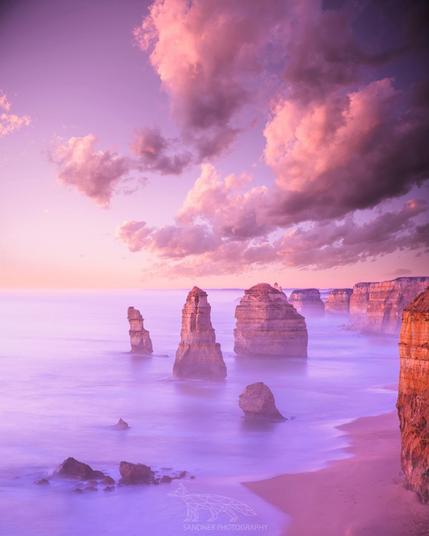A dreamy long-exposure photograph of the Twelve Apostles on Victoria’s Great Ocean Road at twilight. The limestone sea stacks rise dramatically from the smooth, misty ocean, softened by the long exposure. Above, large cotton-candy pink clouds glow in the fading light, casting a serene pastel tone across the entire scene. The golden cliffs on the right frame the coastline, while the foreground rocks and sandy beach add texture and depth to the composition.