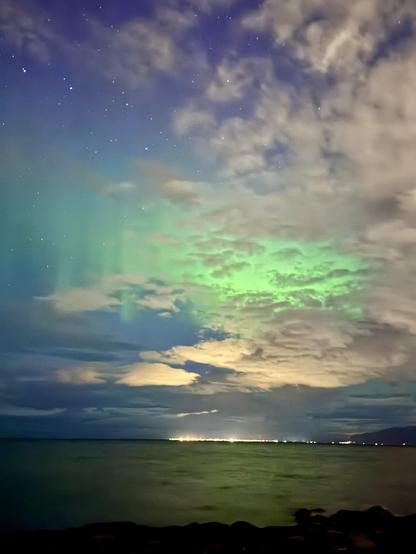 Some green aurora and clouds can be seen over the water with city lights in the distance