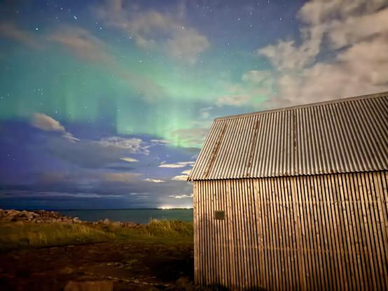 A line of green aurora and clouds above a shack on the side of the road in Seltjarnarnes 