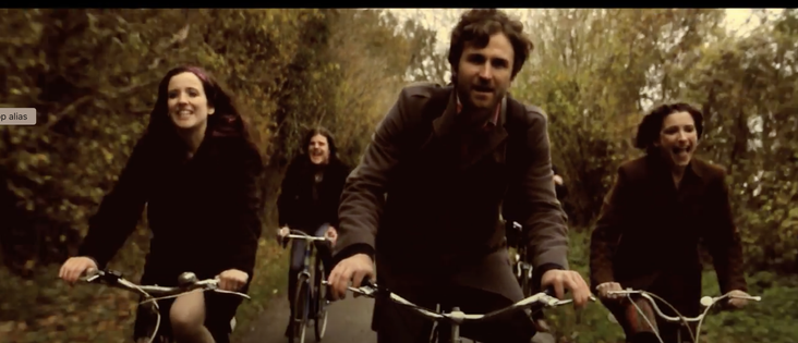 he image depicts four individuals riding bicycles on a narrow path surrounded by trees, suggesting a forest or park setting. The path is lined with dense foliage, and the scene is bathed in a sepia tone, giving it a vintage or nostalgic feel. The individuals are dressed in dark, autumnal clothing, with coats and jackets, indicating a cool climate. The person in the foreground is a man with a beard, wearing a dark jacket, and he is smiling and looking directly at the camera. To his left, a woman…