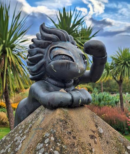 A bronze statue of a lemming from the game Lemmings looking over the top of a pillar. It hangs on to the pyramidal shaped top with its right hand, looking into the distance shielding its eyes with the left. Palm trees and other flowers like in a park or garden are visible behind. Clouds in the sky, but some blue visible as well.