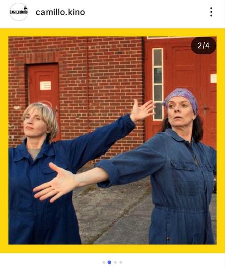 Ein Instagram-Beitrag, der einen Screenshot aus einem Film zeigt. Das Bild hat einen dicken gelben Rand. Im Vordergrund stehen zwei Frauen vor einer roten Backsteinwand mit zwei roten Türen. Beide Frauen tragen blaue Arbeitsanzüge und haben ihre Arme seitlich ausgestreckt, als würden sie tanzen oder eine Choreografie ausführen. Die Frau auf der linken Seite hat kurzes blondes Haar. Die Frau auf der rechten Seite hat dunkle Haare und trägt ein gemustertes lila Bandana. Beide Frauen schauen zur Seite.