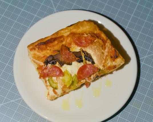 Focaccia pizza corner piece. Toppings include whole milk mozzarella, mushrooms, salami, pepperoncini. An olive oil drizzle is featured on the plate.