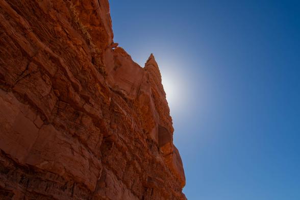 Red sandstone spire rises sharply into a deep blue sky in Capitol Reef National Park, its peak haloed by the midday sun hidden just behind it. Warm light enhances the rock’s texture and stratified layers.