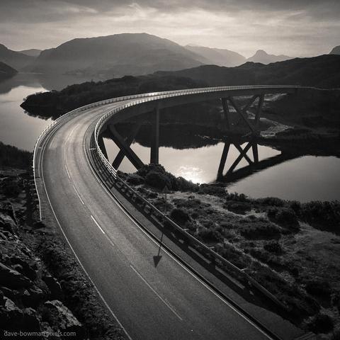 A photograph of the Kylesku Bridge curving in a sweeping arc across calm waters, its reflection mirrored clearly below. The scene is captured in monochrome, with the rugged Highland hills fading softly into the distance, adding depth and contrast to the bridge’s dramatic yet harmonious shape.