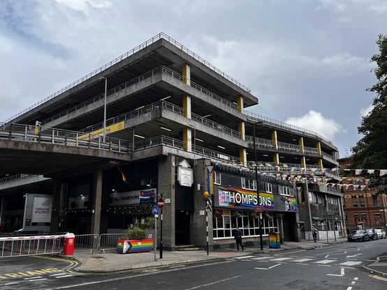 A multistorey car park above a gay pub. There are 4 storeys at the front, with a ramp up to 3 storeys at the back. There's a spiral entrance ramp mostly out of shot to the left. The columns are NCP yellow.