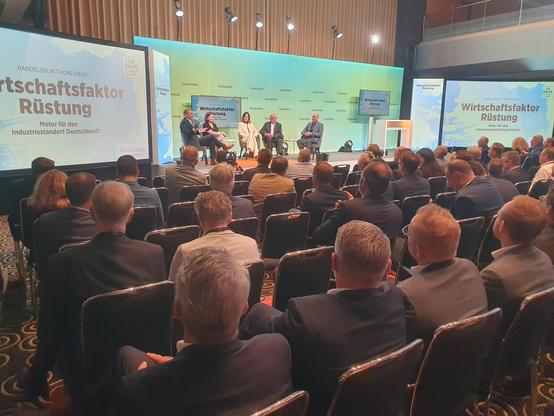 Konferenz mit dem Titel "Wirtschaftsfaktor Rüstung" und dem Untertitel "Motor für den Industriestandort Deutschland?". Auf der Bühne sitzen fünf Personen, die an einem Panel diskutieren. Sie sind in einem großen Saal mit einer großen Anzahl von Zuhörern, die auf Stühlen sitzen und aufmerksam zuhören. Die Bühne ist mit einem grünen Hintergrund und dem Konferenzlogo versehen. Die Zuhörer sind hauptsächlich Männer in Anzügen, was auf eine formelle Veranstaltung hinweist. Die Bildschirme auf beiden…