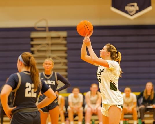 A woman in a white jersey with young hair in a ponytail holds a basketball in two hands as she prepares to the throw it to the left,  another blonde girl in a blue jersey has her arms akimbo as she tracks the ball with her eyes,  we can see another also akimbo in front of her looking away from us with brown hair in a ponytail and the number 22,  and behind them a number of women in white jerseys sitting and looking on with blue folded up bleachers and just a bit of a pennnant with the Ithaca co…