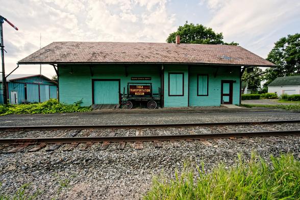 A mostly teal building with a light brown roof and dark brown trim has a sign in front that reads TIOGA TRANSPORTATION MUSEUM with railroad track running from left to right and a little bit of a signal tower at the left,  a matching colored building behind and to the left and some trees and a white building to the right.  There's some grass on the lower right.