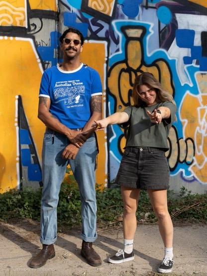 Ein Mann in einem blauen grafischen T-Shirt und Jeans steht neben einer Frau, die ein grünes Hemd und eine schwarze Shorts trägt. Sie posieren spielerisch vor einer bunten Graffiti-Wand, wobei die Frau ein Friedenszeichen hält.