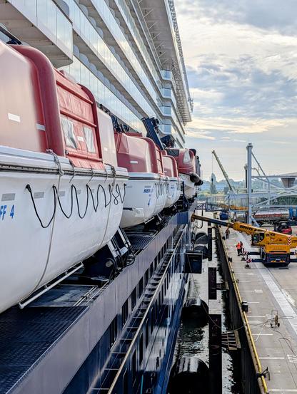 A low-angle, side view of a large cruise ship docked in a port. Several red and white lifeboats are suspended along the side of the ship. In the background, a large yellow crane is visible, along with workers on the dock.