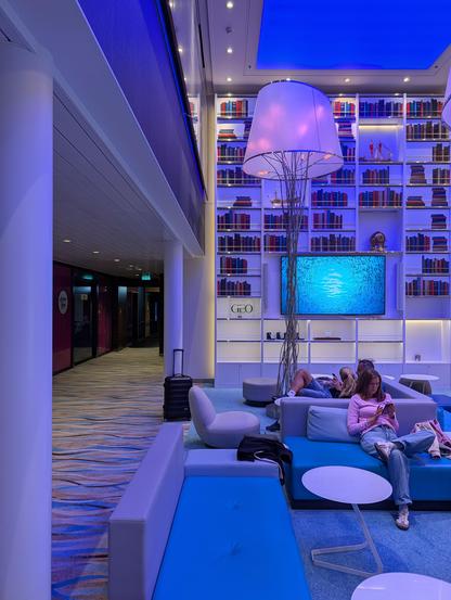 A wide-angle, indoor photo of a modern lounge area on a cruise ship, lit with blue and purple light. There's a wall with white shelves full of books, a large floor lamp, and a TV showing a blue underwater scene. People are sitting on blue and gray sofas in the foreground. A luggage bag is visible next to a chair.