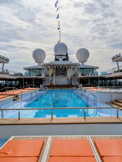 An eye-level, head-on view of the sunny pool deck on a cruise ship. The pool is a vibrant blue, with lounge chairs and seating areas arranged around it. In the background, there is a large bar or stage area with two large white radar domes on top. A series of international flags are flying from a mast.