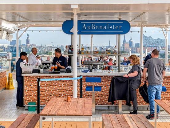 A sunny, wide-angle view of a cruise ship deck bar called "Außenalster," with the city skyline of Hamburg, Germany, in the background. Two bartenders are at the counter, serving a few customers. The deck has wooden tables and benches, and string lights are visible above the bar.