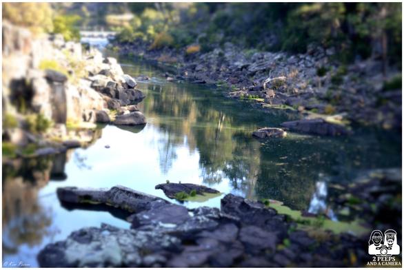  A tilt-shift image of Launceston Gorge in Tasmania, showing a gently flowing river framed by rugged rocks and lush greenery.

The water reflects the trees and sky above, while large boulders scatter across the riverbanks. The tilt-shift effect creates a miniature, dreamlike quality, adding visual intrigue to the natural serenity.

Logo “2 PEEPS AND A CAMERA” appears in the corner, with signature © Kev Peirce.