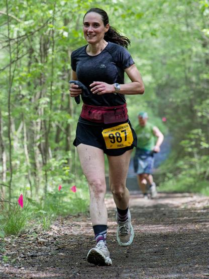 A woman running with a little dirt on her legs on a rather clear dirt trail with a few tiny pink flags on the side,  she's wearing black shorts,  has a yellow bib with the number 987 and a dark grey athletic blouse and has her hair tied back.  Far behind her is a man with blue shorts,  a green shirt and I think a billed cap;  light colored young trees line the side of the trail.
