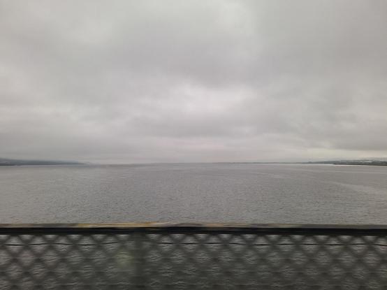 shot from a moving vehicle (train actually) on a bridge, view of a river, sky with grey clouds, water reflecting grey, in the distance to the left and right some low hills are visible.