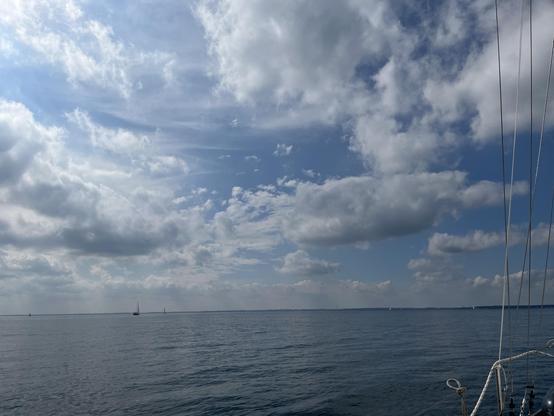 Einige kleine Segelboote vor blauem Himmel mit Wolken. Rechts am Horizont ist etwas Land zu sehen, sonst nur recht glattes Meer.