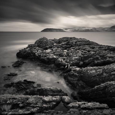 A photograph of the Kintyre peninsula’s rugged shoreline in monochrome, with weathered rocks in the foreground creating texture and depth. Calm waters stretch beyond the rocks toward the distant silhouette of the Isle of Arran, set beneath a dramatic sky. The long exposure softens the movement of clouds and light, contrasting with the solid, rough surfaces of the stone.