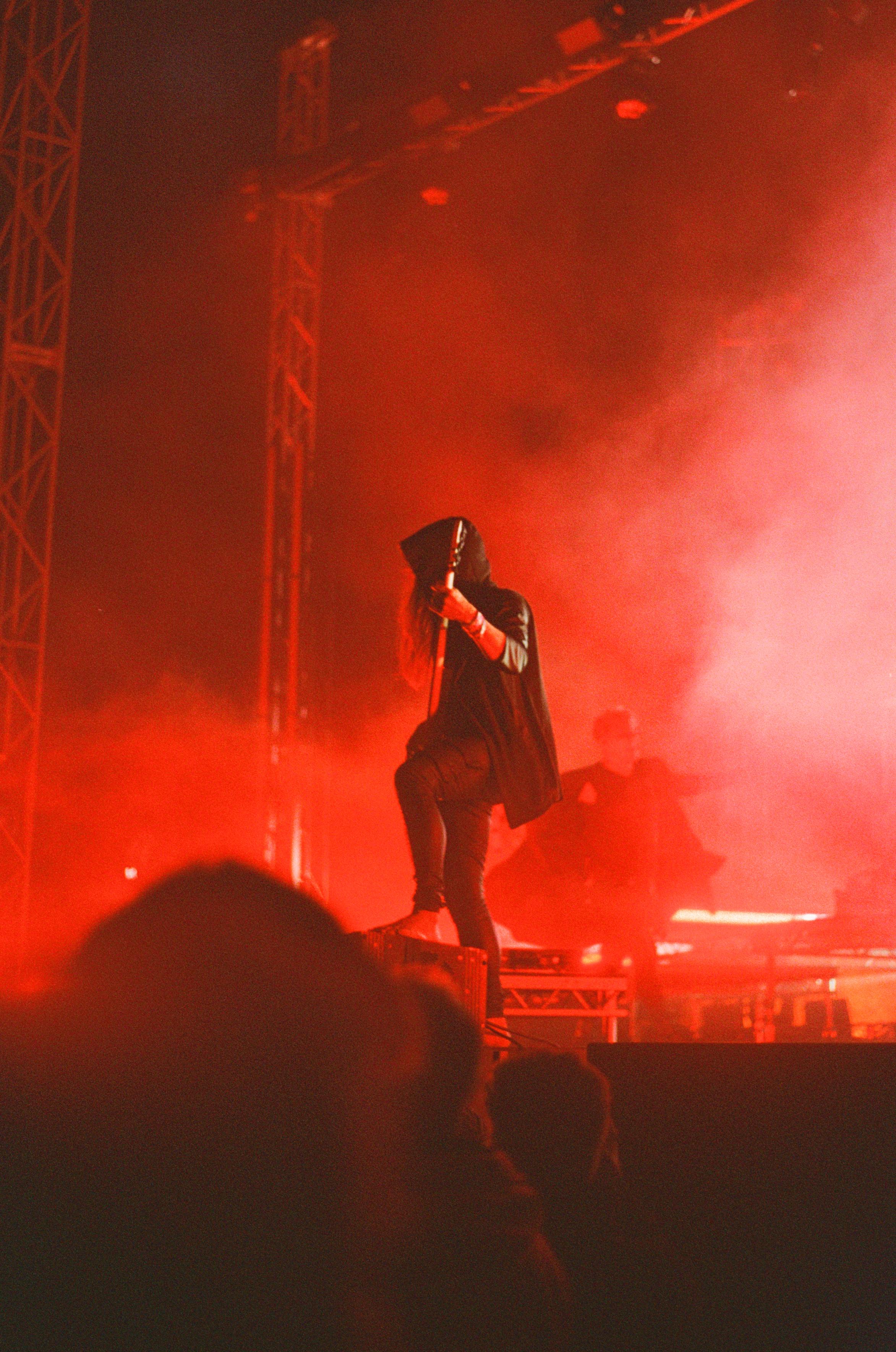 Amos Williams playing bass guitar, dressed in a hoodie, standing at the front of the stage with one leg on an elevation, illuminated by red light.