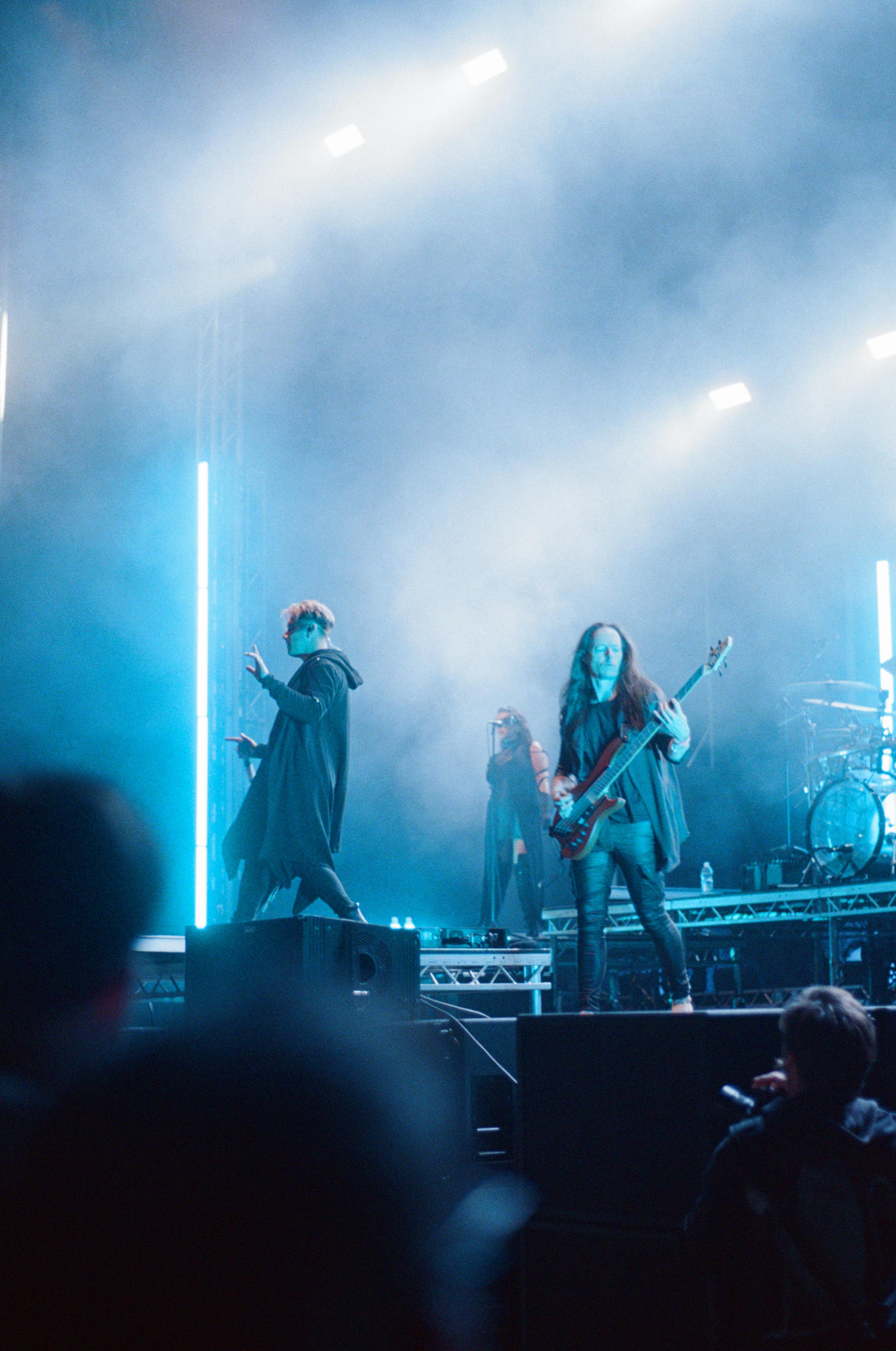 Daniel Tompkins with a microphone in his hand and Amos Williams playing bass guitar, illuminated by light blue light. In the background, a futuristically dressed background vocalist.