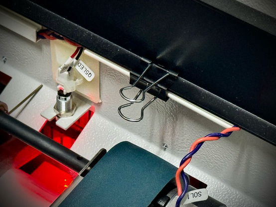 Looking at the innards of printing equipment. A wire that is normally stuck to the frame using a harness with double-sided tape is now clipped to a protruding metal flange using a metal paper binding clip.