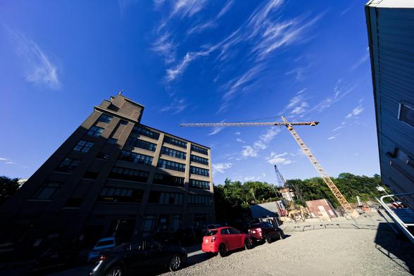 A yellow crane is rotated so the boom is almost horizontal in the frame and below it is a 7 story building which looks small thanks to the wide angle effect,  a row of cars below it is in deep shadow,  to the right can be seen part of a temporary building for the construction site and above most of the frame is full of a blue sky with mainly cirrus clouds.