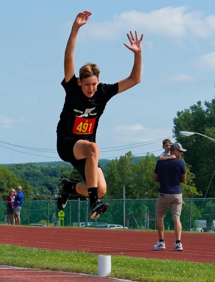 Teenage boy with a black shirt and shorts looks suspended in the air with both arms up and his right leg folded at a 90 degree angle in front of him and the back leg folded back,  behind him on the track there is a man in a billed cap holding a small child,  and a man and a woman hanging out by a fence.
