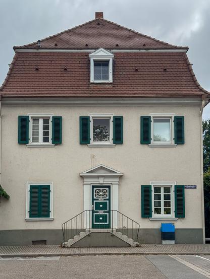 Dreistöckiges Haus mit rotem Mansardealmdach und grünen Fensterläden. Der Eingang ist mit weissen Säulen und einem dreieckigen Sturz gerahmt und symmetrischer Treppenaufgang komplettiert den Blick auf die dreipanelige grüne Tür. Während die unteren 2 Panele mit weissen Vierecken und einem weissen Mittelknauf verziert sind, besteht das oberste Panel aus einem Glasfenster, das ein Spinnennetzmuster aus weißen Holzstegen bildet