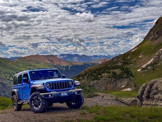Blue Jeep Wrangler Rubicon parked on a rugged mountain trail with sweeping views of the San Juan Mountains under a partly cloudy summer sky.